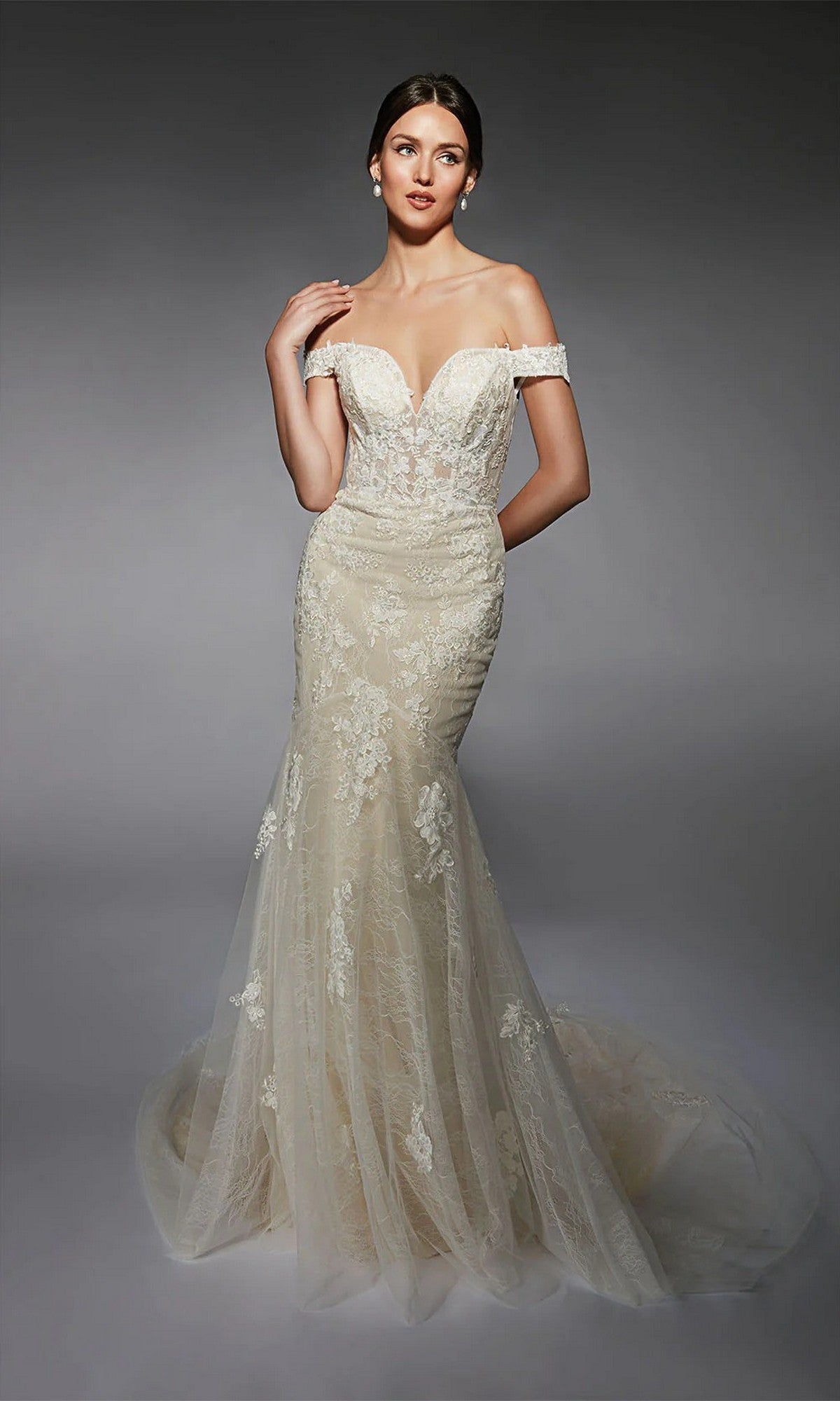 A woman poses against a dark background in the Alyce 7118 long white dress, an ivory/champagne off-the-shoulder gown with lace details, a dramatic mermaid silhouette, and a sweeping train for timeless elegance.